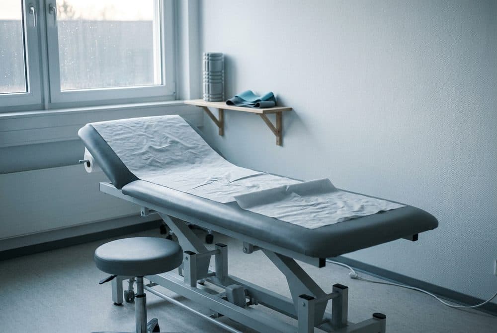 Sala de atendimento de fisioterapia com maca, banquinho e luz natural entrando pela janela